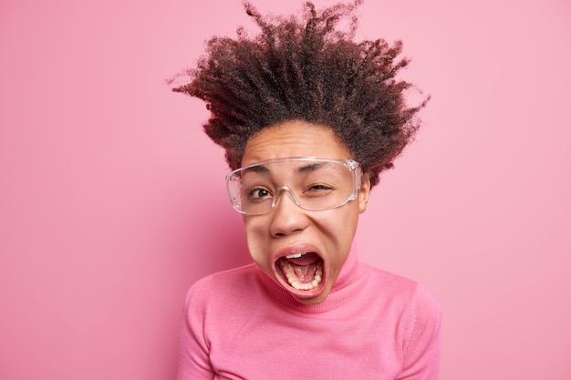 Studio shot di una donna afroamericana emotiva dai capelli ricci pazza strizza l'occhio occhio tiene la bocca aperta esclama ad alta voce indossa occhiali trasparenti dolcevita