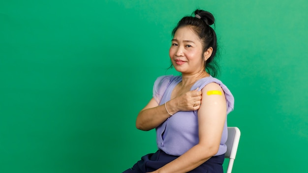 Studio shot di una donna asiatica di mezza età si siede sorridendo e mostra una benda di gesso gialla sul braccio dopo aver ricevuto la vaccinazione contro il coronavirus covid 19 dal medico in clinica su sfondo verde.