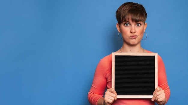 Studio shot di una giovane donna con un'espressione scettica in possesso di una lavagna vuota su sfondo blu. Copia spazio, spazio per il tuo annuncio o testo.