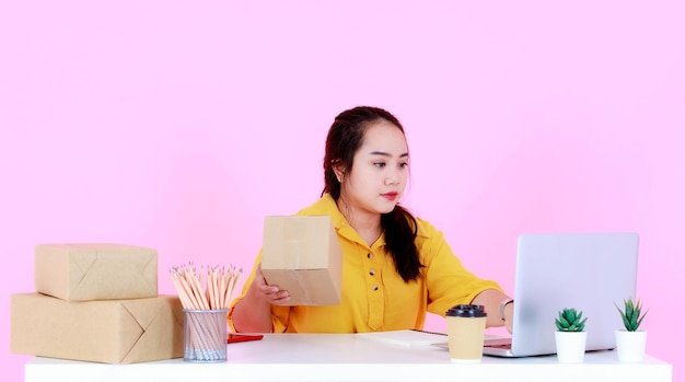 Studio shot di una giovane donna d'affari asiatica di successo startup imprenditrice che controlla l'indirizzo di spedizione della consegna per le scatole del pacchetto di spedizione dal computer portatile sulla scrivania su sfondo rosa.