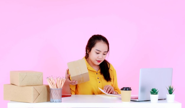 Studio shot di una giovane donna d'affari asiatica di successo startup imprenditrice che controlla l'indirizzo di spedizione della consegna per le scatole del pacchetto di spedizione dal computer portatile sulla scrivania su sfondo rosa.