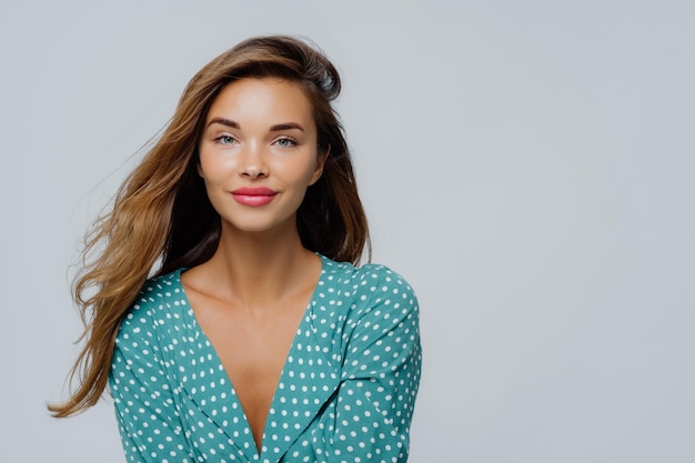 Studio shot di una giovane donna positiva ha lunghi capelli ondulati, il trucco indossa una camicetta a pois turchese guarda direttamente i modelli della fotocamera su sfondo bianco copia spazio per il tuo annuncio