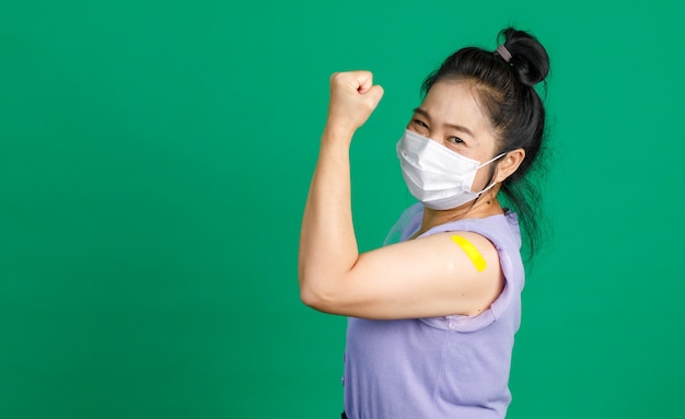 Studio shot di una paziente asiatica di mezza età, felice e sana, indossa una maschera facciale e guarda la telecamera che tiene il pugno alzato mostra i muscoli e l'intonaco della fasciatura blu dopo aver ricevuto il vaccino contro il coronavirus covid 19.