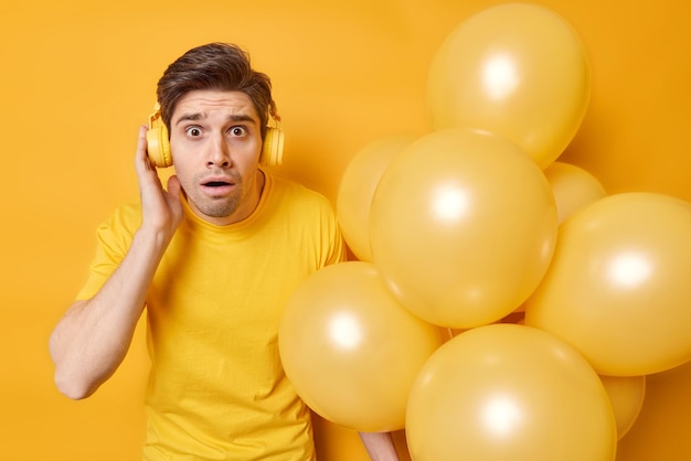 Stupefatto giovane adulto ascolta la musica tramite le cuffie fissa gli occhi infastiditi posa con un mazzo di palloncini gonfiati si prepara per la festa di compleanno isolato su sfondo giallo Concetto di vacanza