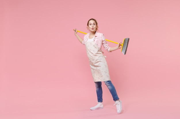 Stupita bella giovane donna casalinga in abiti casual, grembiule che fa i lavori domestici isolati su sfondo rosa pastello parete ritratto in studio. Concetto di pulizia. Mock up spazio di copia. Tenere in mano la scopa.