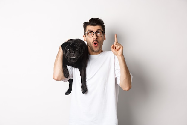 Stupito bell'uomo con gli occhiali, che tiene in mano un simpatico cagnolino nero sulla spalla, puntando il dito verso il logo promozionale, in piedi su sfondo bianco