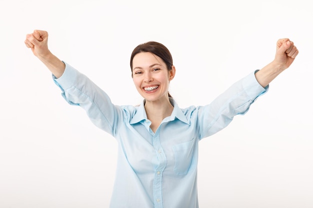 Stupito bruna giovane imprenditrice in camicia casual sta gesticolando la vittoria con le mani alzate è scioccata estremamente felice sorriso raggiante su sfondo bianco