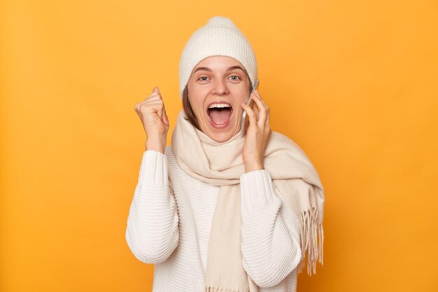 Stupito estremamente felice donna caucasica in piedi in caldo berretto maglione e sciarpa parlando su smart phone sentendo notizie eccellenti pugno chiuso celebrando la gioia isolato su sfondo giallo