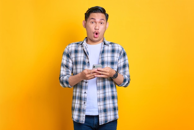 Stupito giovane asiatico in camicia a quadri utilizzando il telefono cellulare e guardando la fotocamera isolata su sfondo giallo