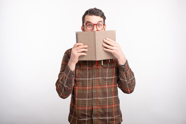 Stupito giovane con gli occhiali su spazi bianchi si copre il viso con un libro aperto. Momento della lettura.