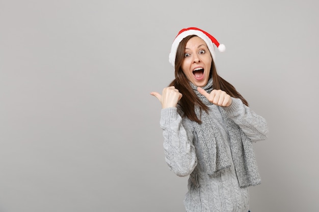 Stupito giovane Santa ragazza in maglione grigio sciarpa cappello di Natale tenendo la bocca spalancata, puntando i pollici da parte urlando isolato su sfondo grigio. Felice Anno Nuovo 2019 celebrazione festa concetto.