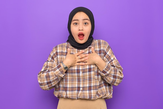 Stupito la giovane donna asiatica in camicia a quadri con le mani sul petto guardando la fotocamera con l'espressione del viso wow isolata su sfondo viola