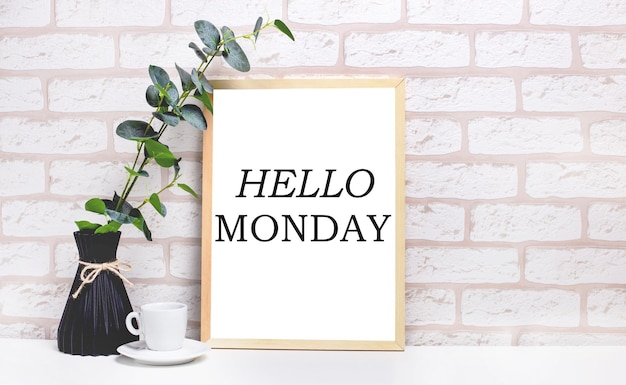 Su un tavolo bianco contro un muro di mattoni chiari, un ramo di eucalipto in un vaso scuro, una tazza bianca e una cornice di legno chiaro con il testo HELLO MONDAY. Interno del Ministero degli Interni.