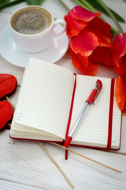 Su un tavolo di legno bianco c'è un quaderno rosso con tulipani, c'è una tazza di caffè e un pan di zenzero a forma di labbra scarlatte