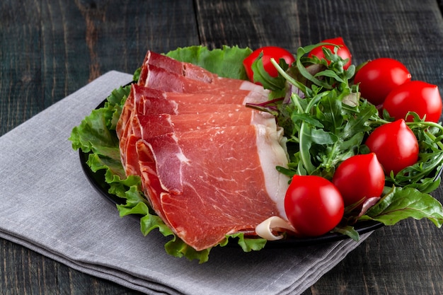 Su un tavolo di legno c'è un piatto con prosciutto a fette, pomodori ed erbe aromatiche