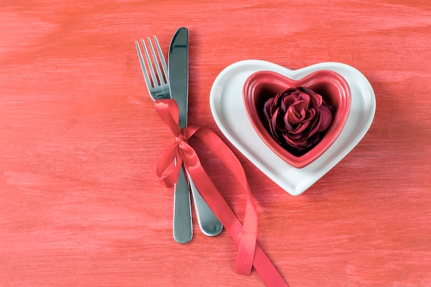 Su un tavolo rosso due piatti a forma di cuore, un bocciolo di rosa rossa e posate legati con un nastro rosso