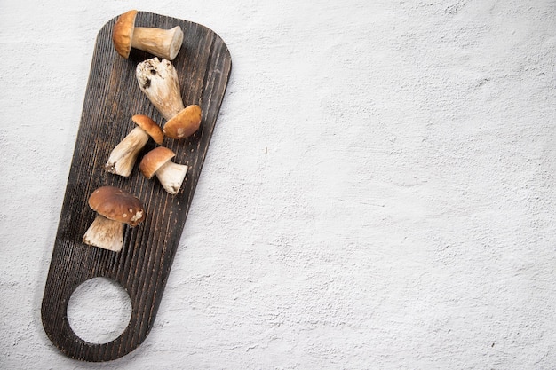 Su uno sfondo chiaro si trova un tagliere di legno scuro su cui sono presenti i funghi porcini di bosco