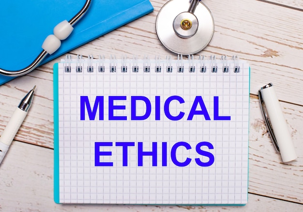 Su uno sfondo di legno chiaro c'è uno stetoscopio, un quaderno blu, una penna bianca e un foglio di carta con la scritta MEDICAL ETHICS. Concetto medico