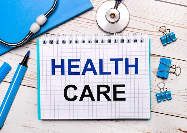 Su uno sfondo di legno chiaro, uno stetoscopio, un blocco note blu, graffette blu, un pennarello blu e un foglio di carta con il testo HEALTH CARE. Concetto medico