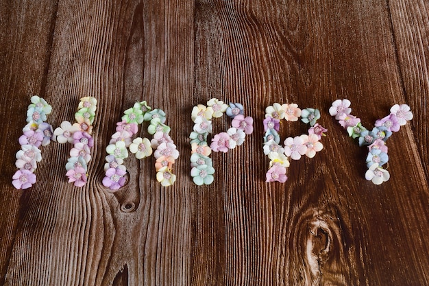 Su uno sfondo di legno scuro, la parola HAPPY è allineata con fiori multicolori a grandi lettere.