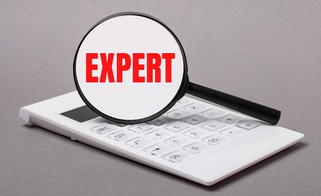 Su uno sfondo grigio una calcolatrice bianca e una lente d'ingrandimento con il testo EXPERT Business concept
