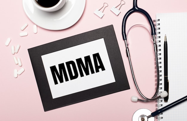 Su uno sfondo rosa chiaro, un taccuino con una penna, uno stetoscopio, pillole bianche, graffette e un foglio di carta con il testo MDMA