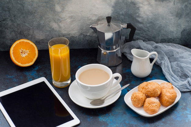 su uno sfondo scuro caffè con latte, torte, caffettiera, tavoletta, garza come decorazione e un bicchiere con succo d&#39;arancia