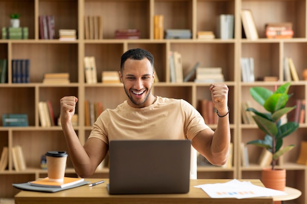 Successo aziendale Uomo arabo felicissimo che usa il laptop e gesticola sì agitando i pugni celebrando la grande fortuna