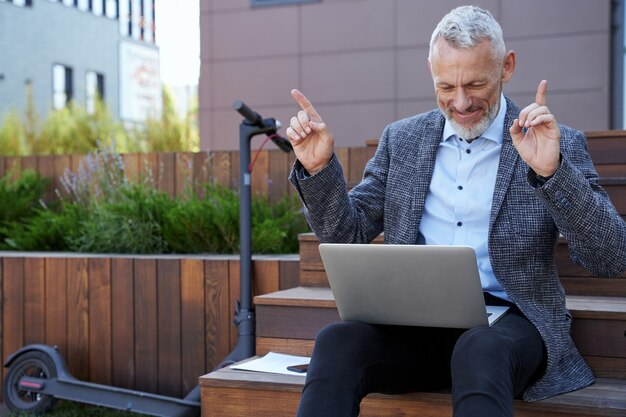 Successo felice uomo d'affari maturo che ha un ottimo umore mentre si lavora al laptop seduto all'aperto su