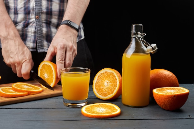 Succo d'arancia fresco in un bicchiere e una bottiglia con frutta fresca affettata con un coltello. L'uomo taglia le arance al tavolo di legno su sfondo nero