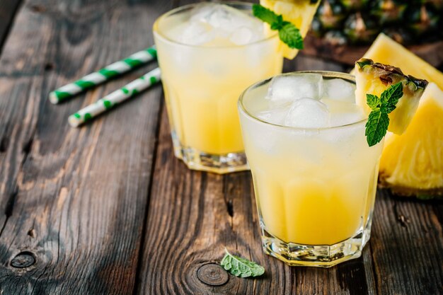 Succo di ananas fresco in due bicchieri con ghiaccio e foglie di menta su fondo rustico scuro