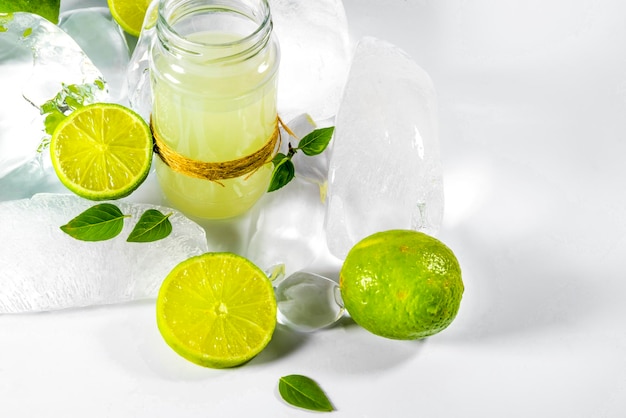 Succo di lime nel bicchiere, fette di lime, foglie di basilico e ghiaccio su sfondo bianco.