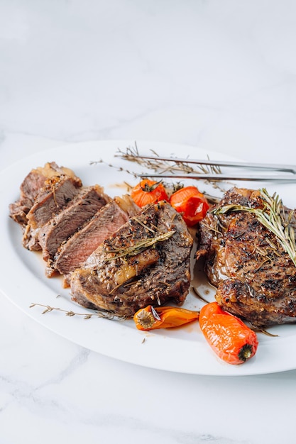 Succose bistecche di ribeye ben fritte su un piatto bianco su una superficie di marmo bianco con lampone e pomodorini e peperoni fritti Le bistecche sono affettate e pronte per essere servite