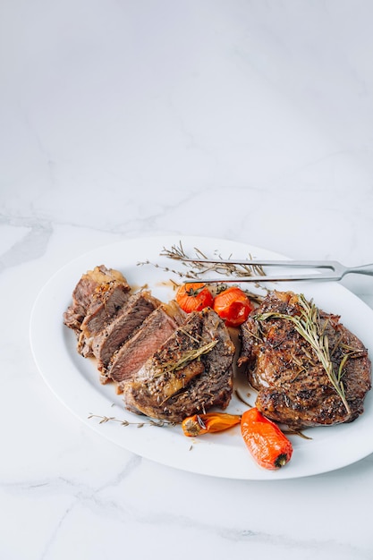 Succose bistecche di ribeye ben fritte su un piatto bianco su una superficie di marmo bianco con lampone e pomodorini e peperoni fritti Le bistecche sono affettate e pronte per essere servite