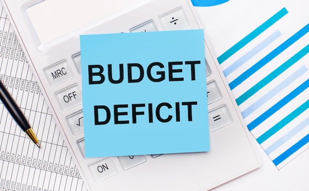 Sul desktop c'è una calcolatrice bianca con un adesivo blu con il testo BUDGET DEFICIT, una penna e dei report blu. Concetto di affari