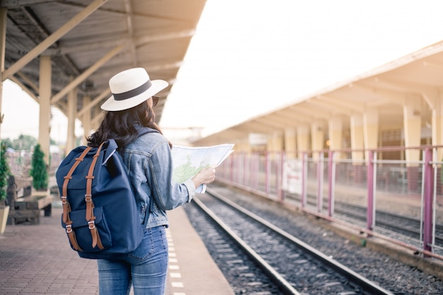 Sul retro di donne asiatiche che portano una borsa con gli occhiali neri, indossa un cappello e tiene in mano una mappa alla stazione ferroviaria.