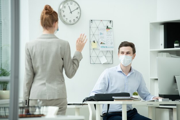 Sul retro di una giovane donna d'affari elegante che agita la mano al suo collega maschio in maschera protettiva seduto alla scrivania davanti al computer in ufficio