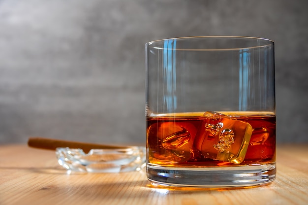 Sul tavolo c'è un bicchiere di whisky con cubetti di ghiaccio. Un posacenere con un sigaro sfocato