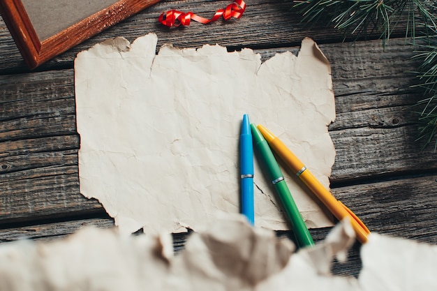 Sul tavolo di legno ci sono una vecchia carta gialla che dice: Caro Babbo Natale rosso, matite, penne, colori assortiti regalo e rami di albero per il nuovo anno