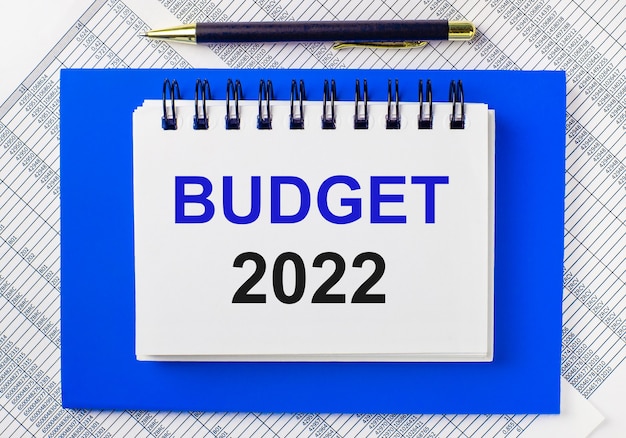 Sullo sfondo dei rapporti sul desktop, un blocco note blu. Ha una penna e un taccuino bianco con il testo BUDGET 2022. Concetto di business