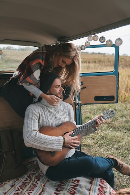 Suona la musica che le piace. Bel giovane che suona la chitarra per la sua bella ragazza mentre è seduto in mini van in stile retrò