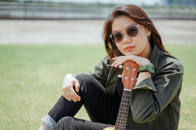 Suonare l'ukulele di giovane bella donna asiatica che indossa giacca e jeans neri in posa all'aperto