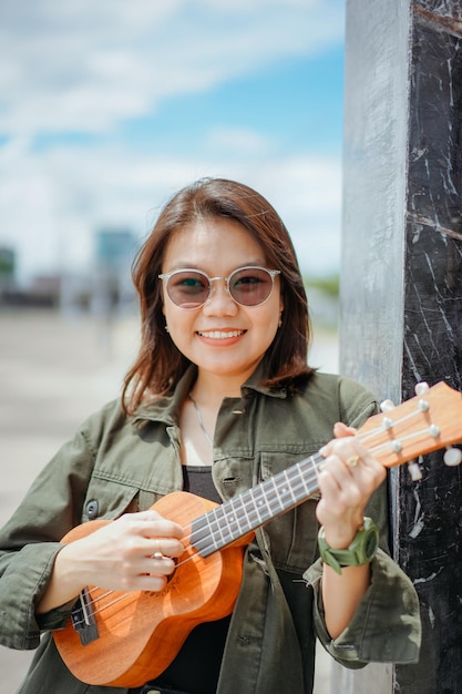 Suonare l'ukulele di giovane bella donna asiatica che indossa giacca e jeans neri in posa all'aperto