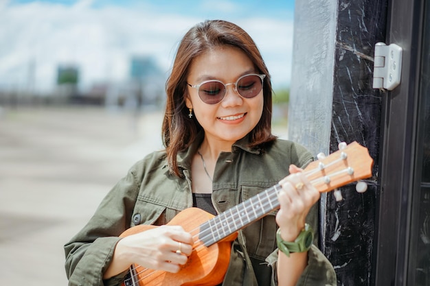 Suonare l'ukulele di giovane bella donna asiatica che indossa giacca e jeans neri in posa all'aperto