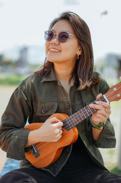 Suonare l'ukulele di giovane bella donna asiatica che indossa giacca e jeans neri in posa all'aperto