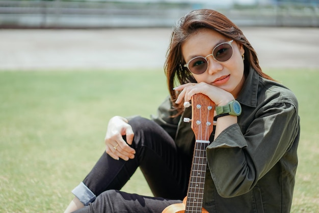 Suonare l'ukulele di giovane bella donna asiatica che indossa giacca e jeans neri in posa all'aperto