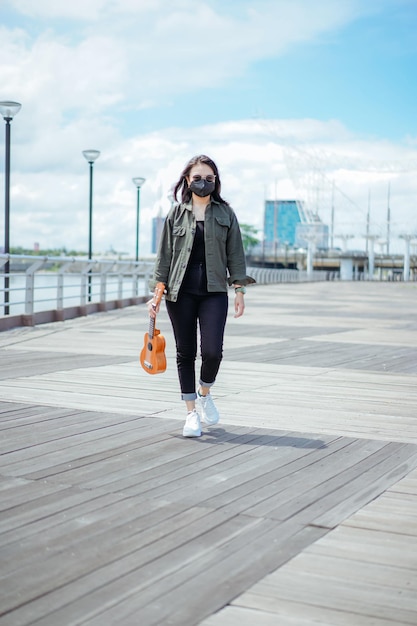 Suonare l'ukulele di giovane bella donna asiatica che indossa giacca e jeans neri in posa all'aperto