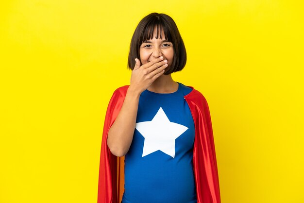 Super eroe donna incinta isolata su sfondo giallo felice e sorridente che copre la bocca con la mano