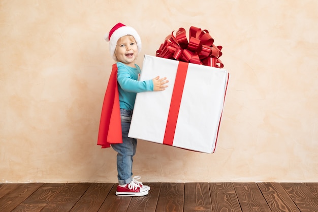 Supereroe vestito bambino felice che tiene il contenitore di regalo di Natale. Bambino divertente che indossa un costume da super eroe con un regalo di Natale