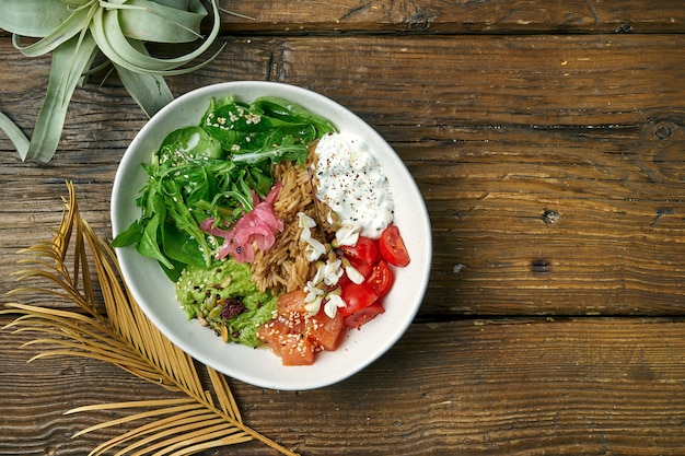 Superfood - una ciotola con riso, salmone, guacamole, uovo in camicia e pomodorini conditi con yogurt greco su un legno. Cibo equilibrato